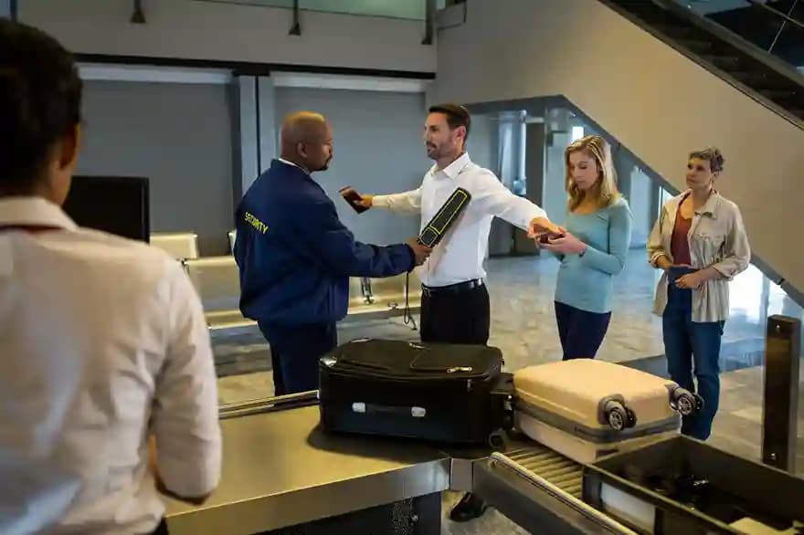 An airport security officer screening a traveler with a handheld detector beside a luggage belt