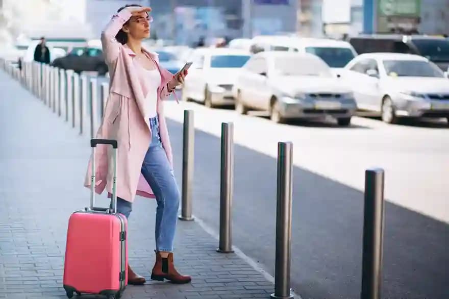 A traveler waiting at the curb with a suitcase, shading her eyes as she watches for her vehicle